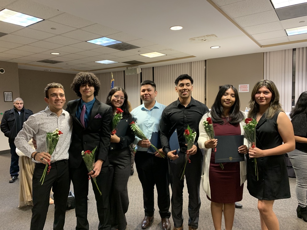 7 Graduates standing shoulder to shoulder each holding a flower.