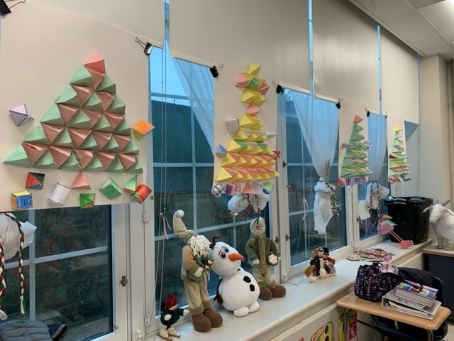 Two green and 2 yellow holiday trees made using geometric patters hung on the white shades of the windows with stuffed characters standing in the window sill